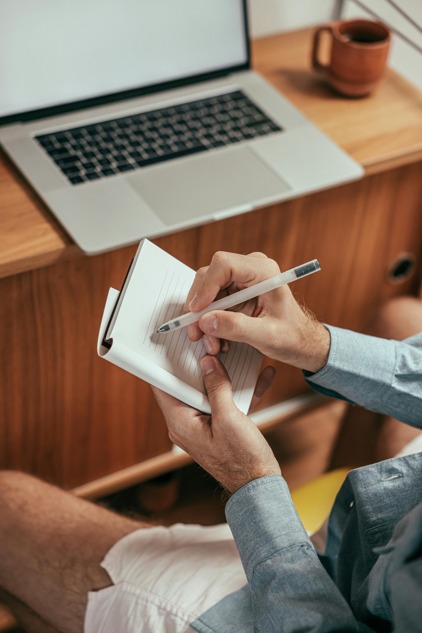 A person writing notes in a small pad beside a laptop.